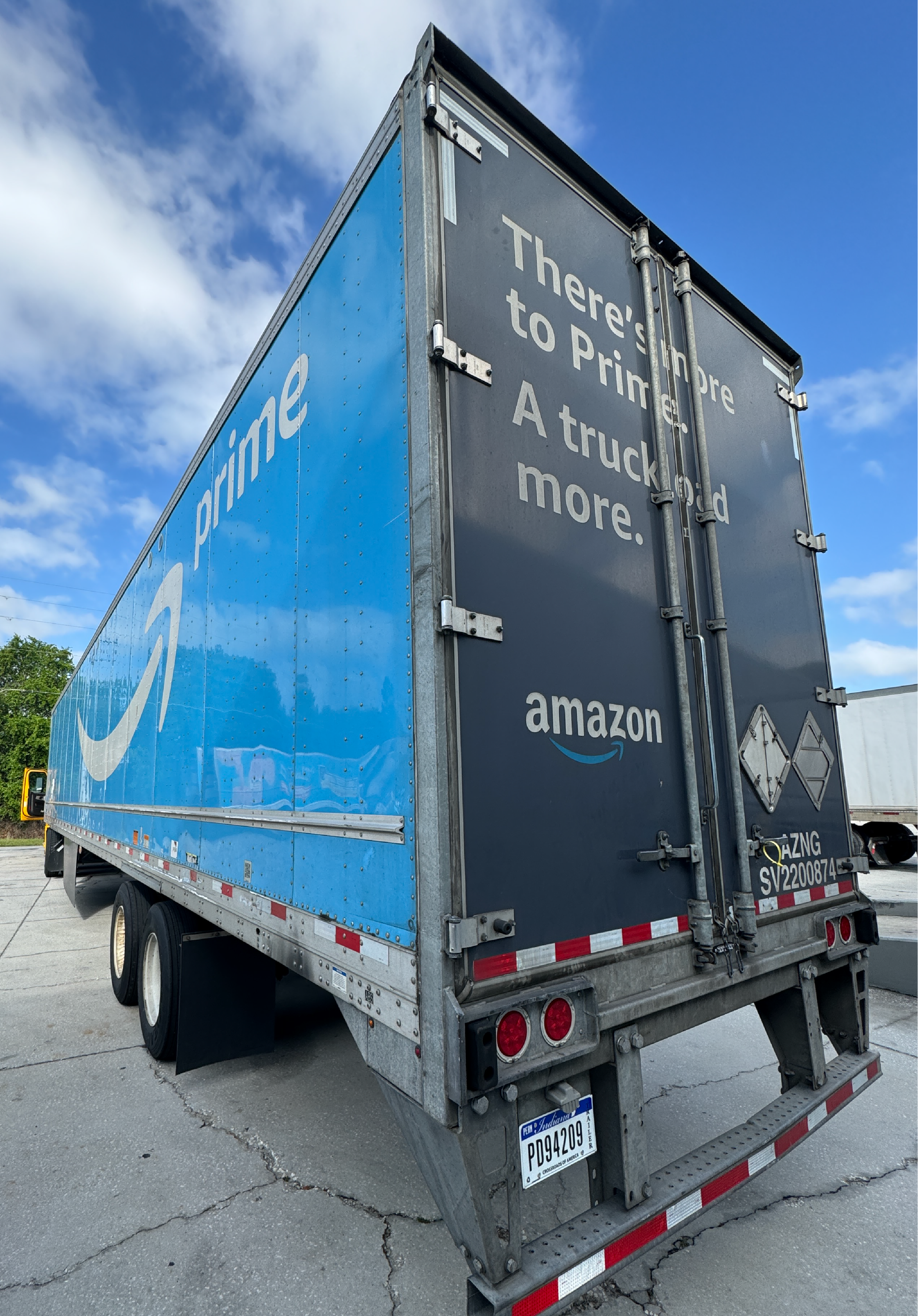Camión de entrega de Amazon Prime con el eslogan 'There's more to Prime. A truckload more.' estacionado sobre el pavimento bajo un cielo azul con nubes.