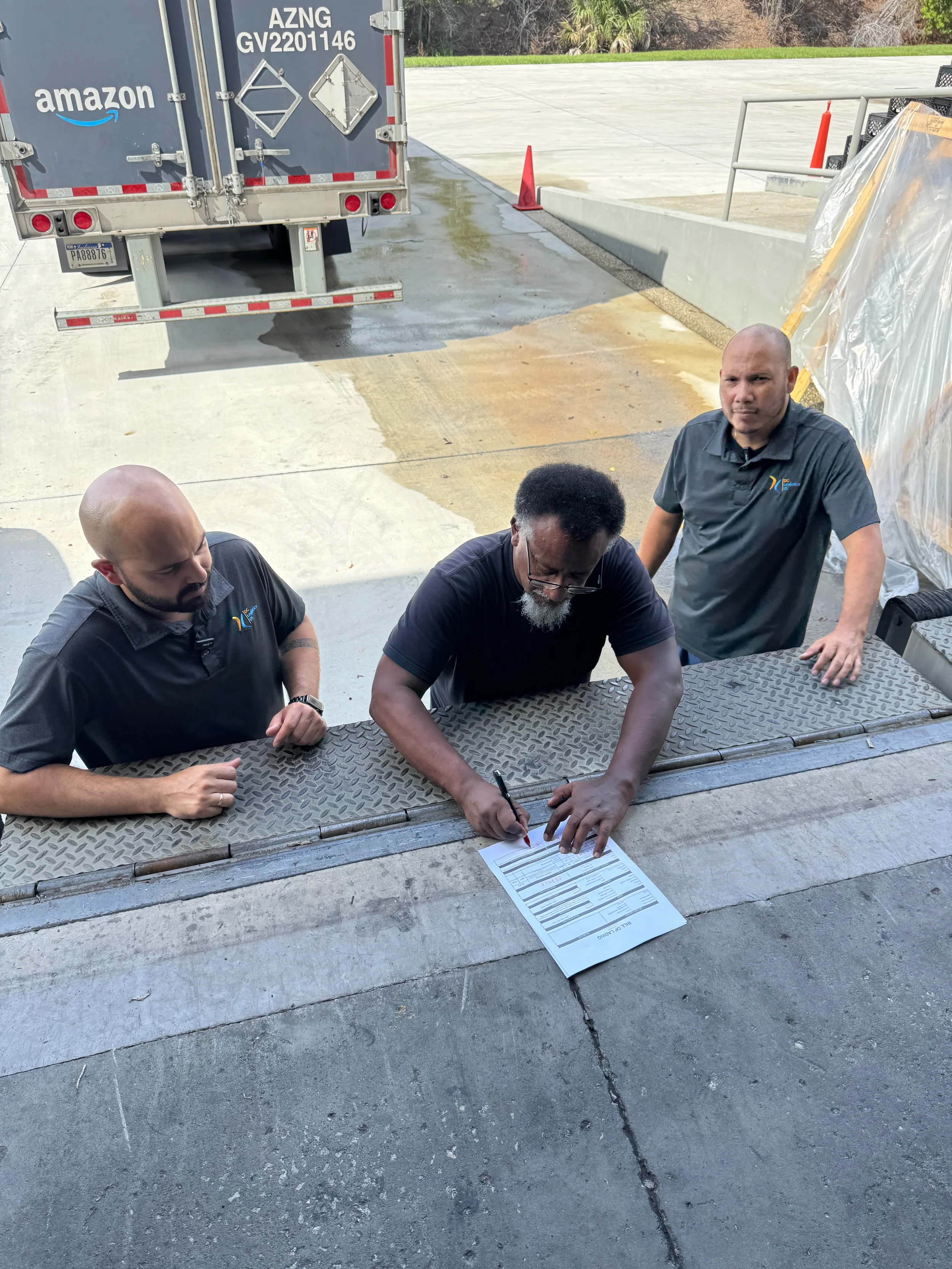 Tres hombres están reunidos alrededor de una mesa al aire libre, uno de ellos firmando un documento mientras los otros dos observan. Detrás de ellos hay un camión y algunos materiales de construcción.