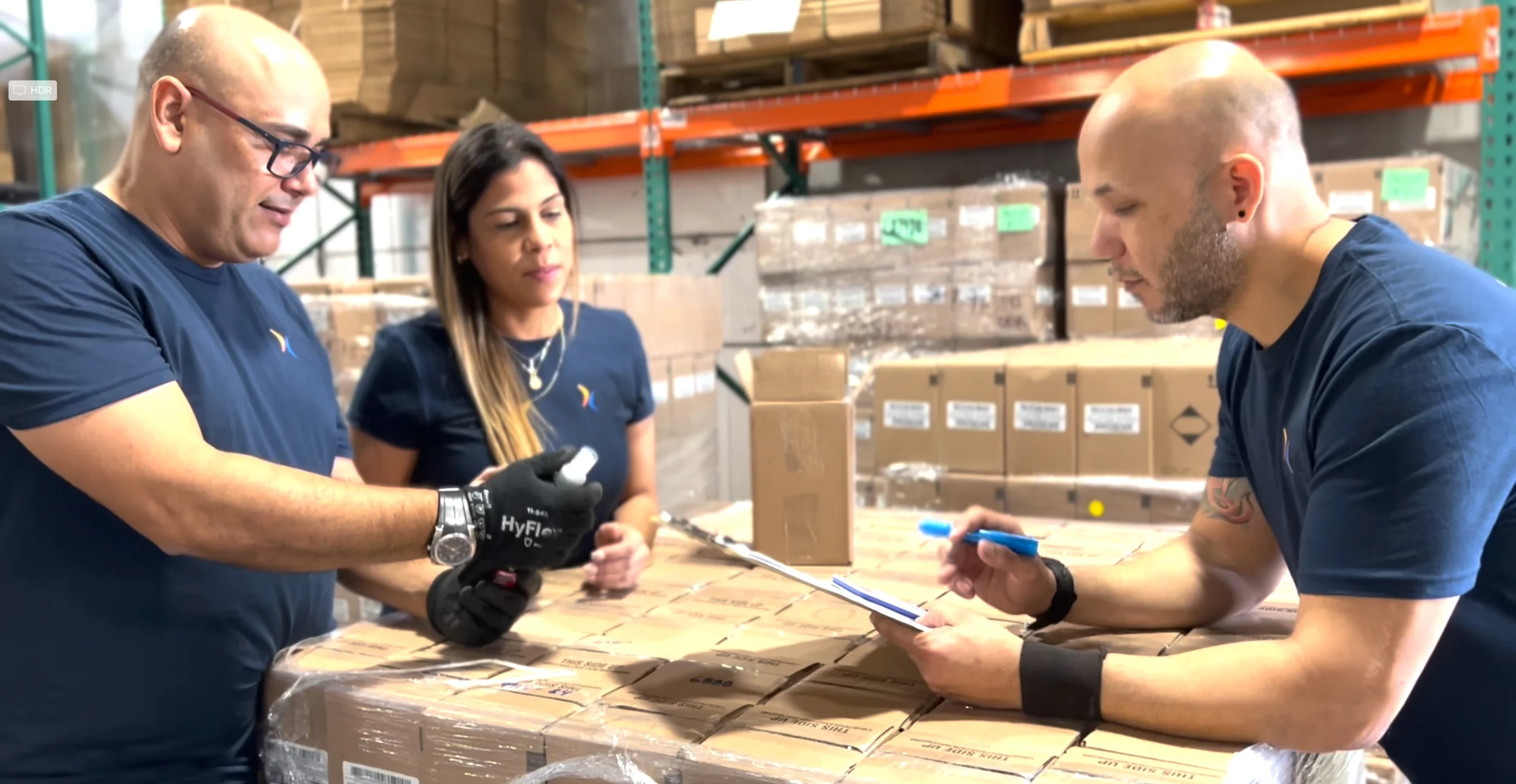 Tres personas trabajando juntas en un almacén, inspeccionando y registrando información sobre cajas de suministros en un palet.