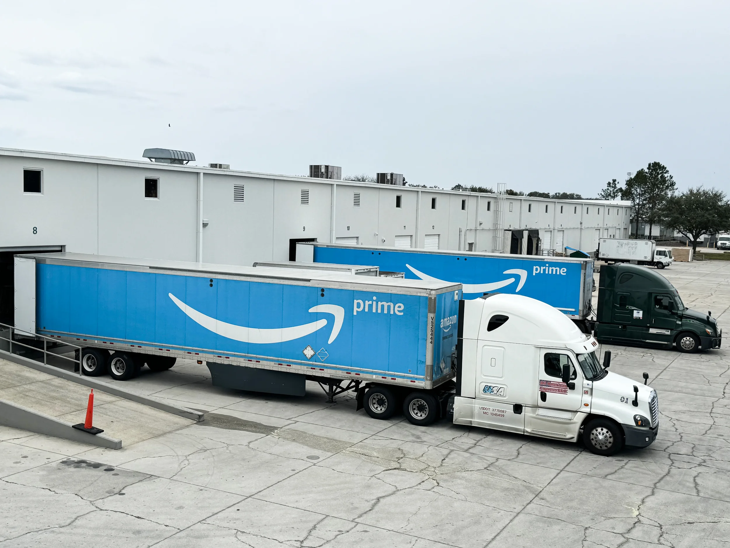 Un camión semirremolque con la marca de Amazon Prime estacionado en un patio cerca de un gran almacén blanco.