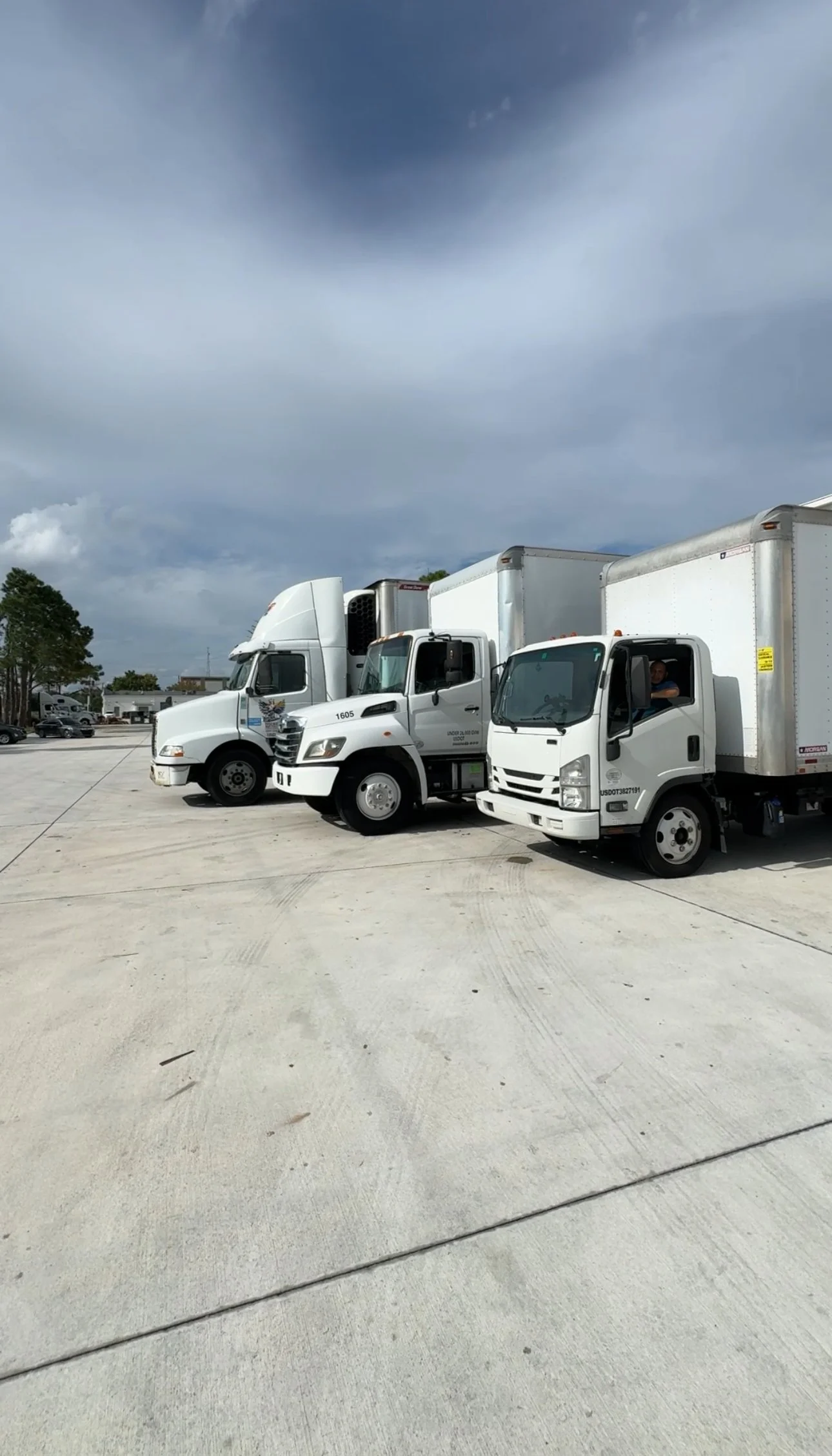 Tres camiones de reparto blancos estacionados en un amplio lote de concreto bajo un cielo mayormente nublado.