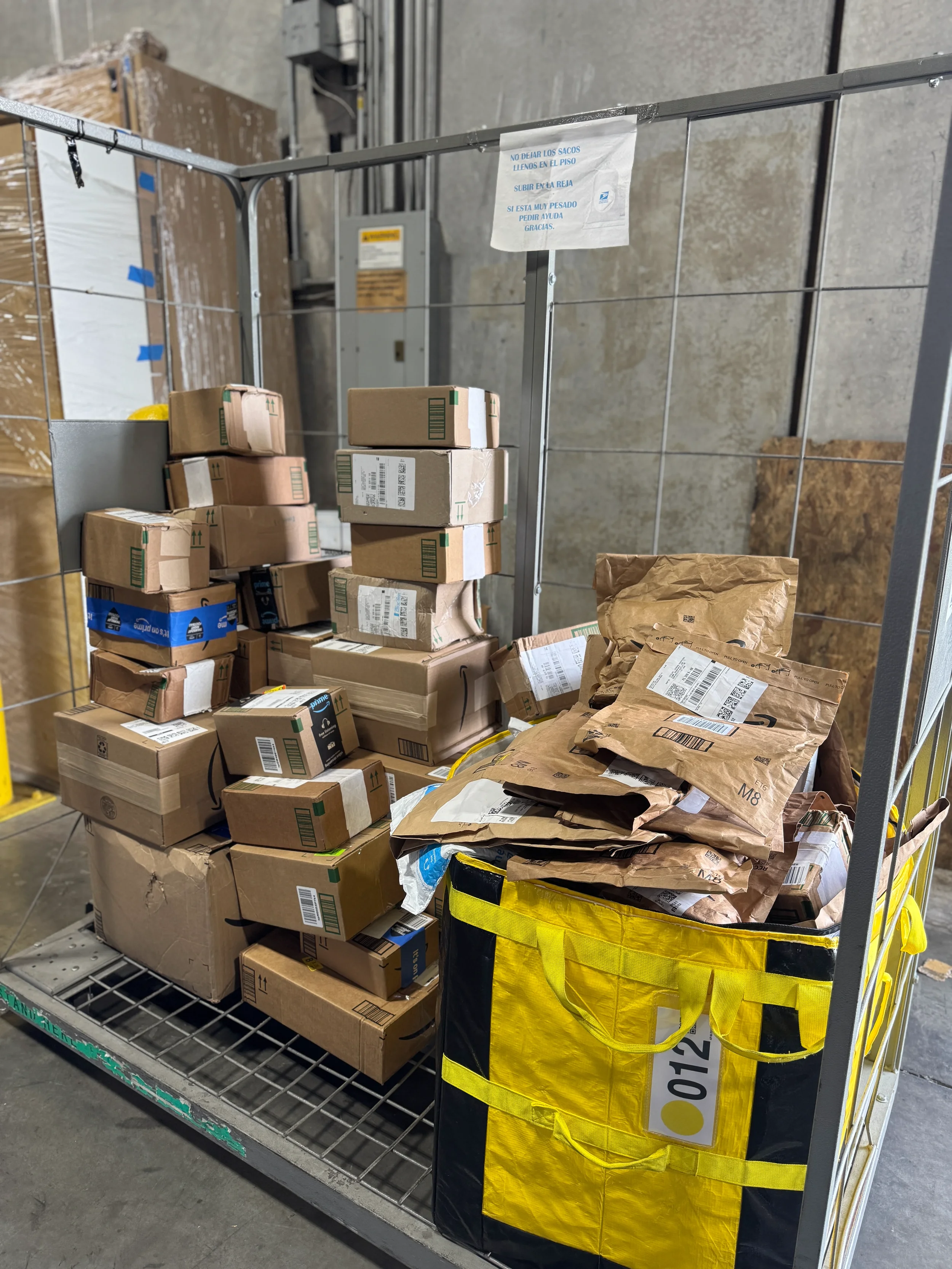 Un carrito de metal lleno de diversas cajas y paquetes de papel marrón, algunos con etiquetas de envío, en un entorno de almacén.