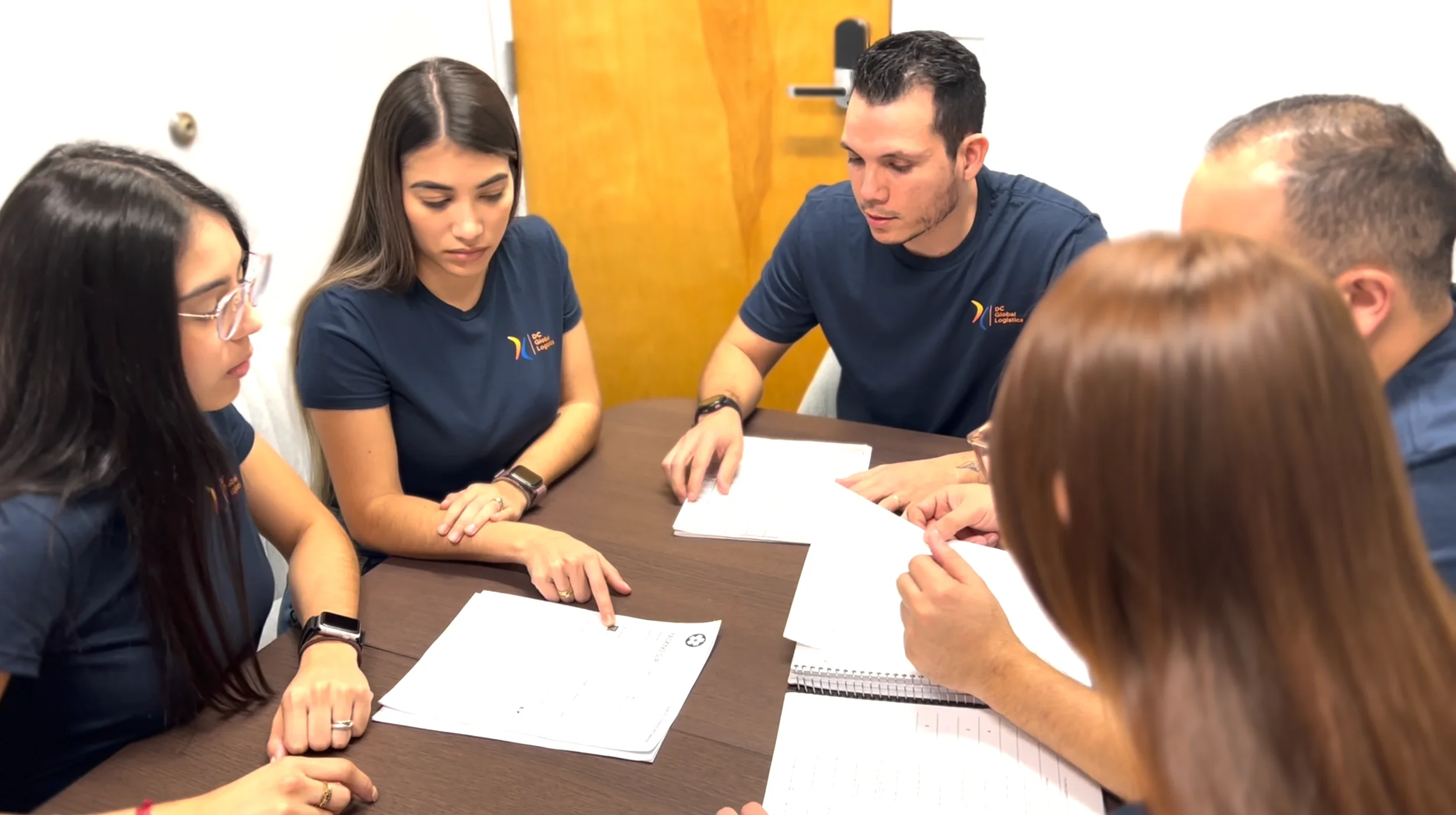 Seis personas sentadas alrededor de una mesa de conferencias, participando en una conversación con documentos impresos y cuadernos, vistiendo camisas azul marino con un logo.
