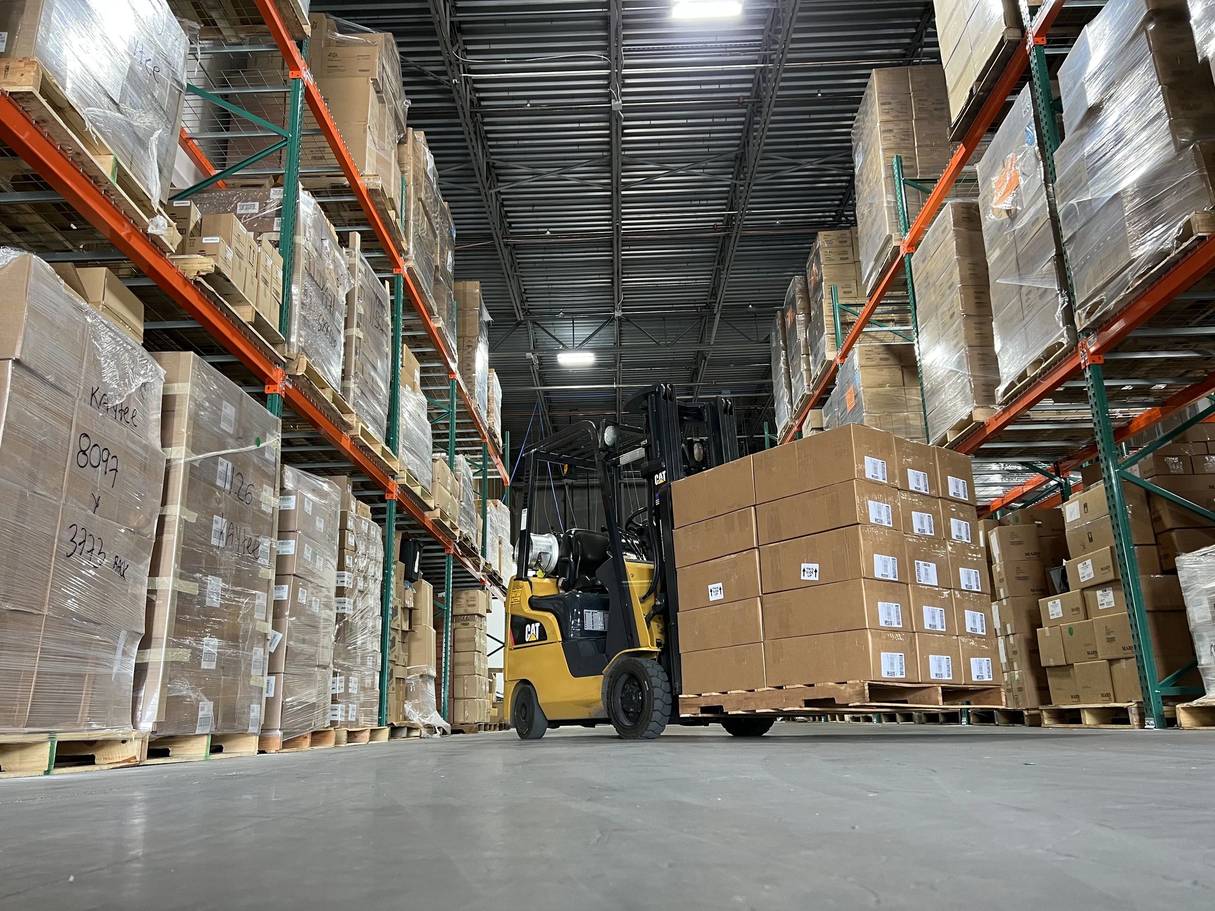 Un montacargas Caterpillar amarillo moviendo un palet de cajas de cartón en un almacén con estanterías altas surtidas de cajas.