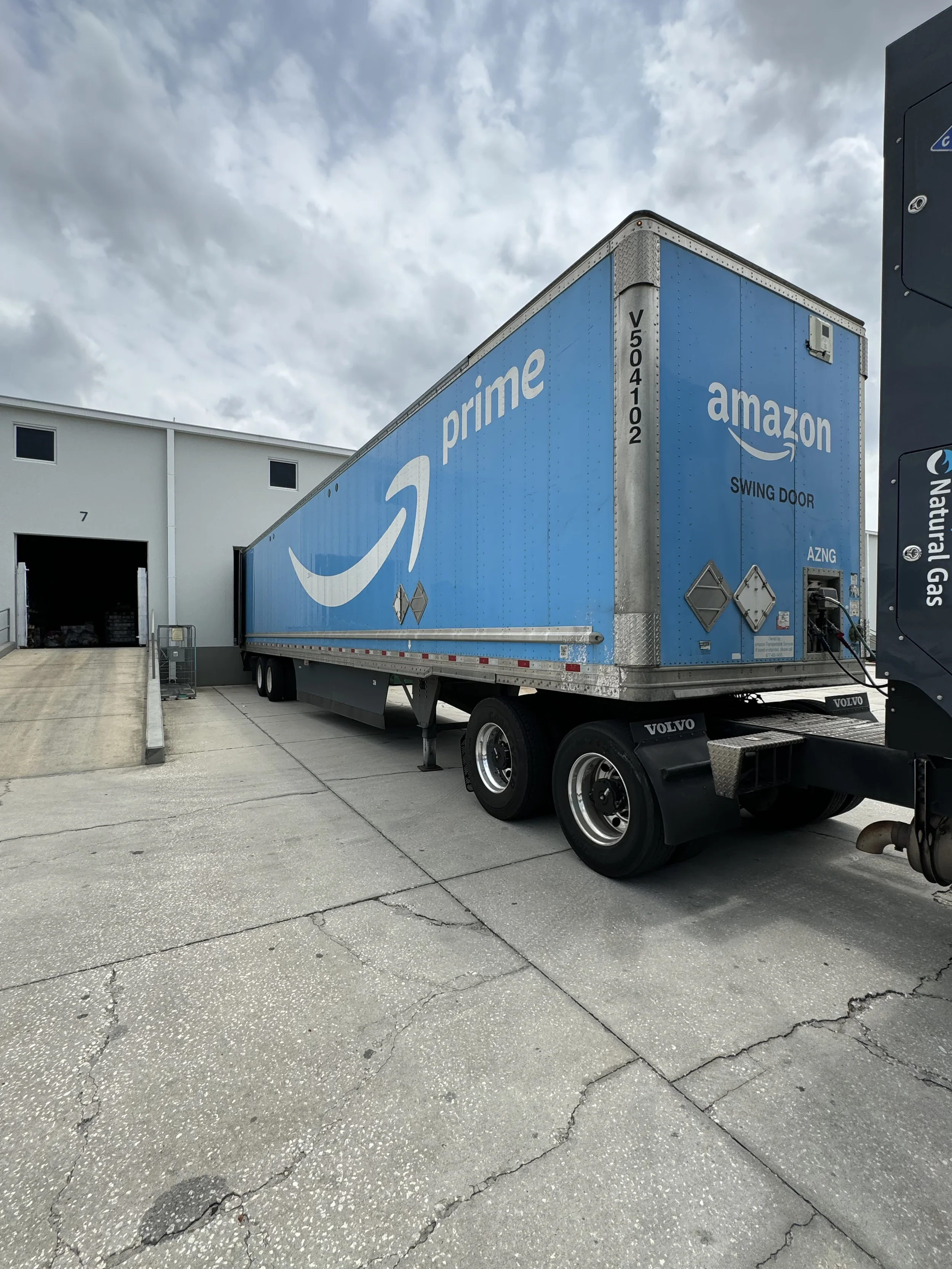 Camión azul de Amazon Prime con puerta abatible estacionado afuera de un almacén bajo un cielo nublado.