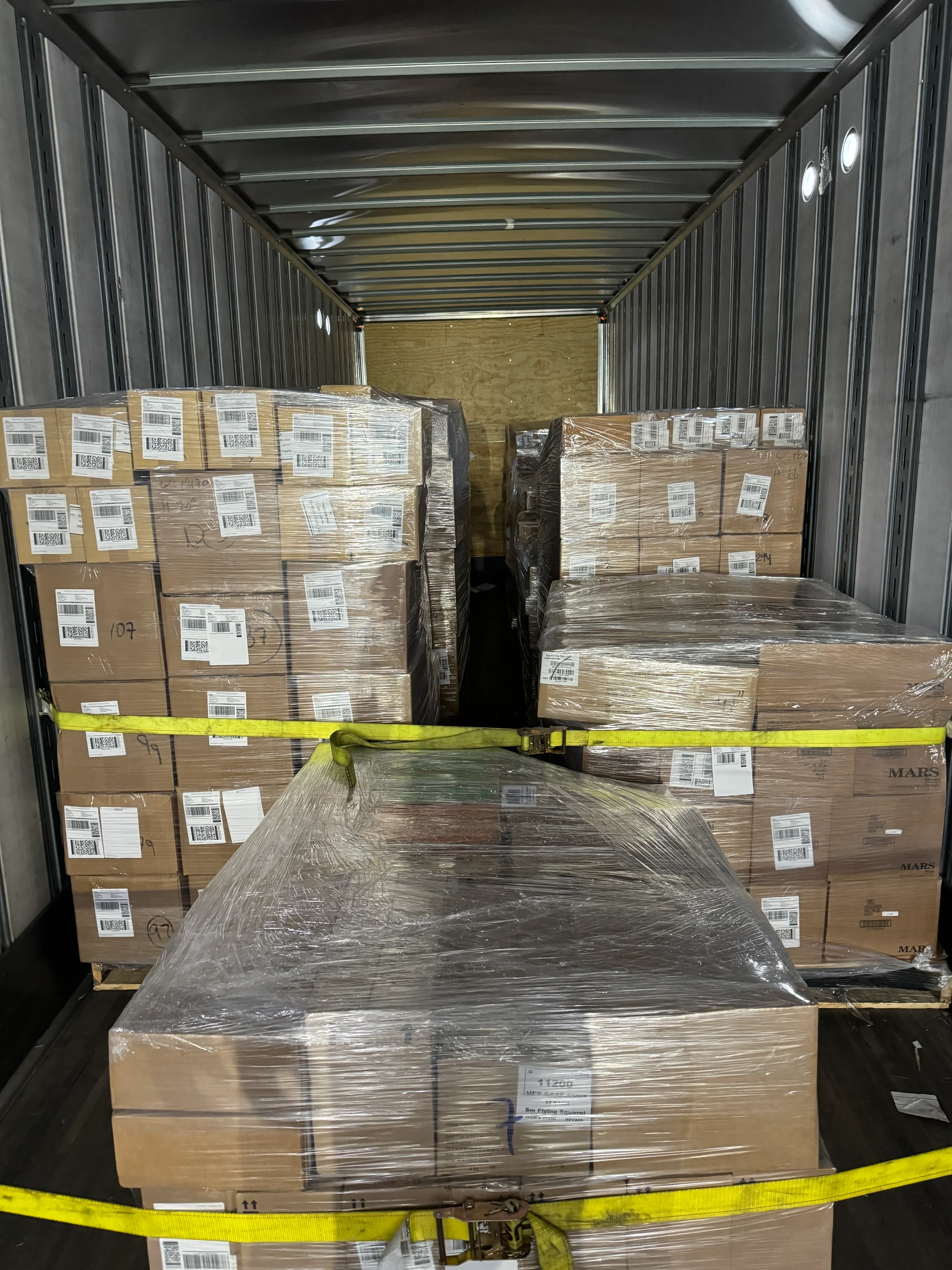Interior de un remolque de camión lleno de cajas de cartón apiladas envueltas en plástico, aseguradas con correas amarillas.