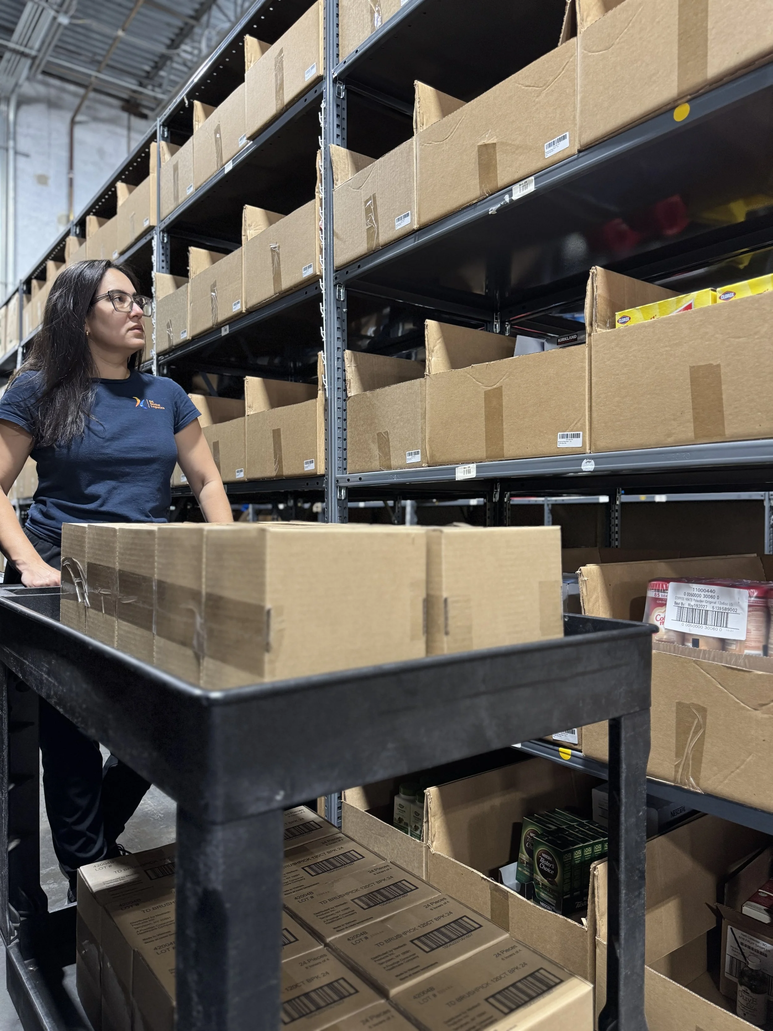 Una mujer con cabello largo y oscuro y gafas, vestida con una camisa azul marino, está de pie junto a un carrito negro lleno de cajas dentro de un almacén con estanterías industriales que sostienen más cajas.