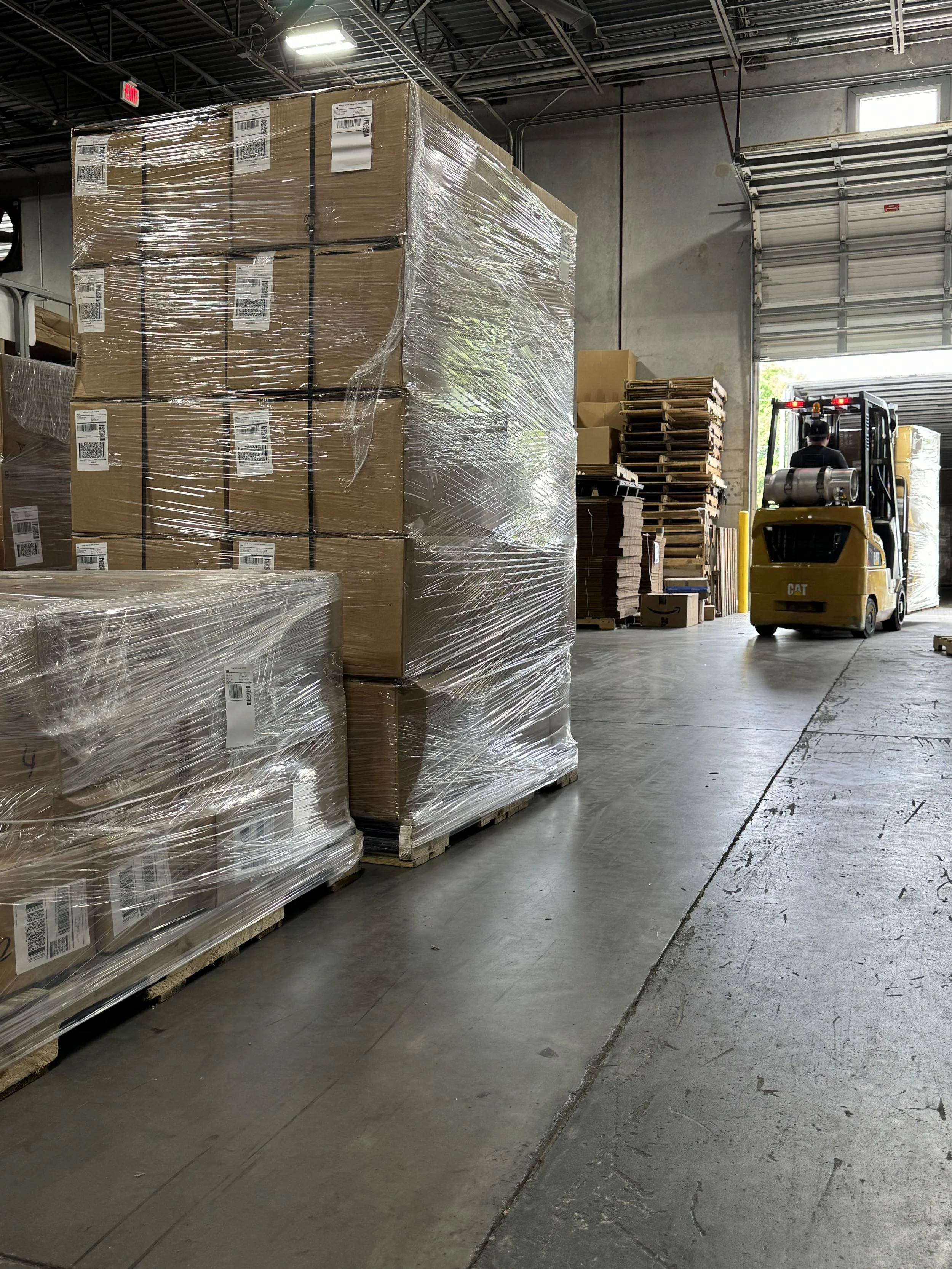 Palets de cajas de cartón envueltas en plástico en un almacén industrial, con un montacargas amarillo y cajas apiladas vistas cerca de una puerta de garaje parcialmente abierta.