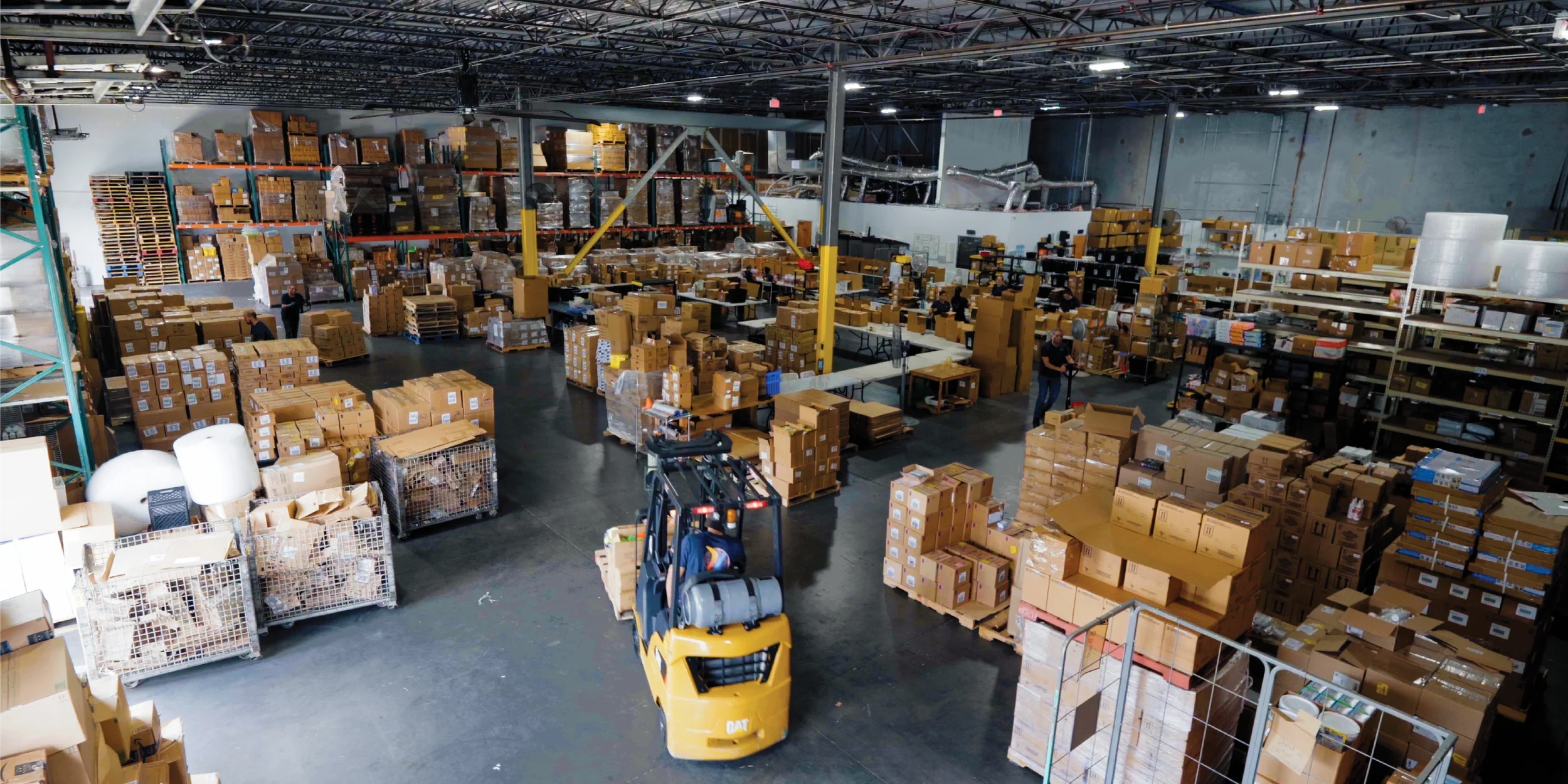 Almacén con estanterías de cajas, trabajadores y un montacargas moviendo palets.