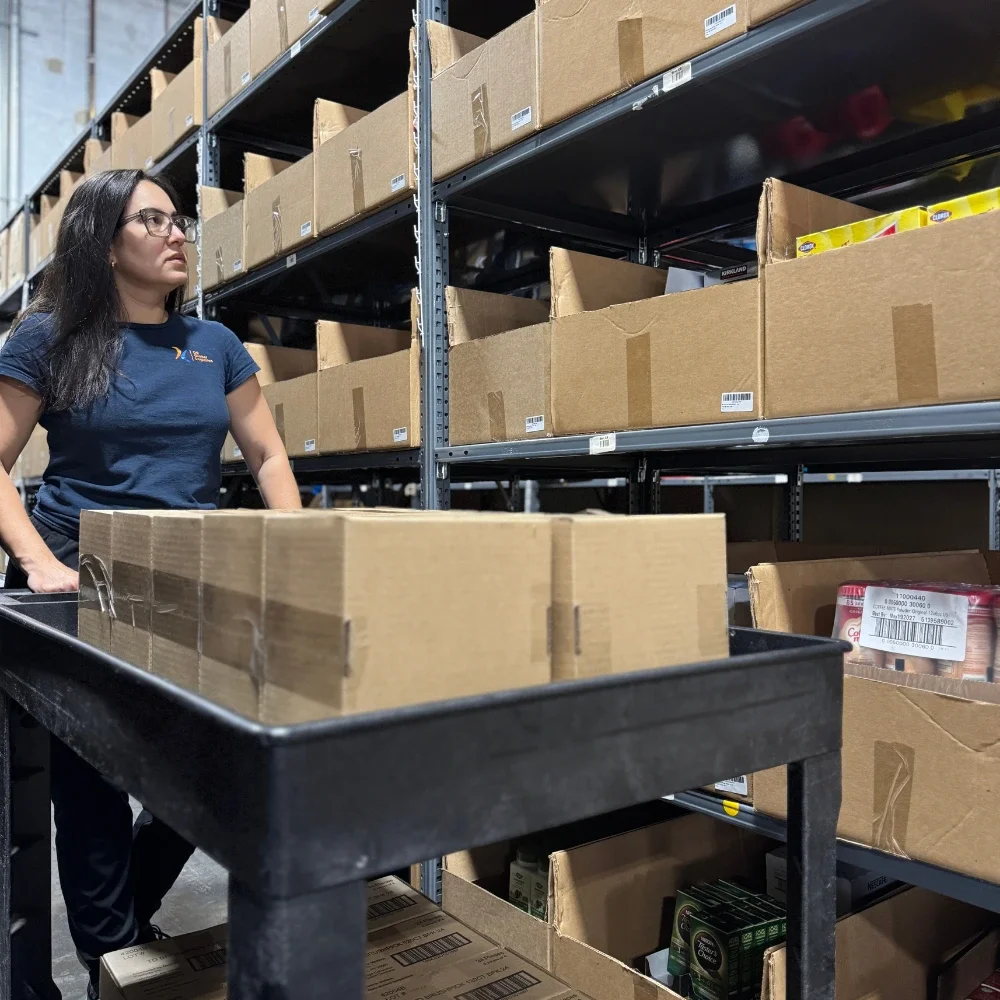 Una mujer con gafas y una camisa azul marino de pie en un pasillo de almacén con estanterías llenas de cajas de cartón y productos, junto a un carrito negro con cajas de cartón vacías apiladas.