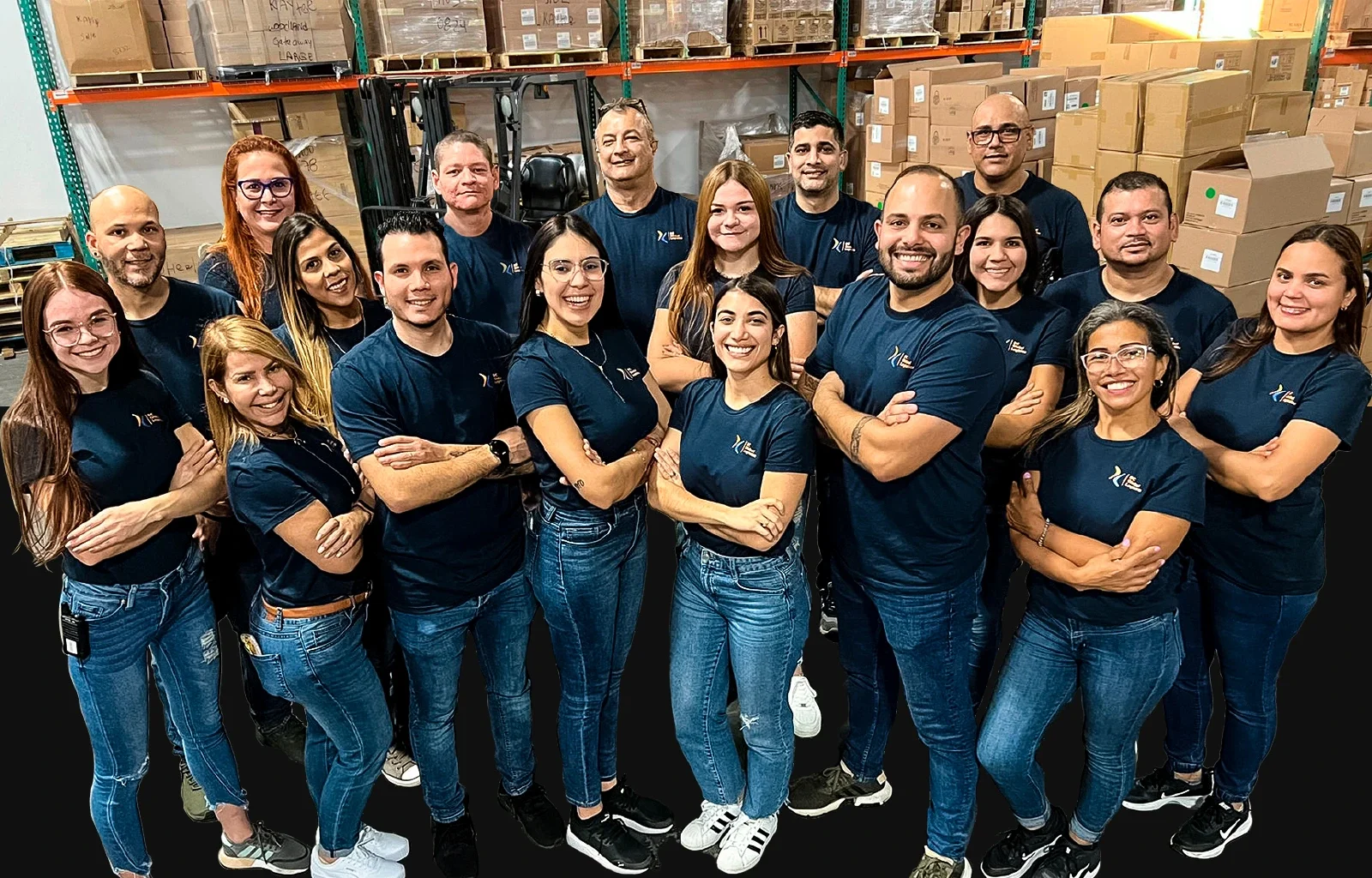 Grupo de 17 personas de pie en un almacén, con camisas azules a juego con un logo, sonriendo a la cámara.
