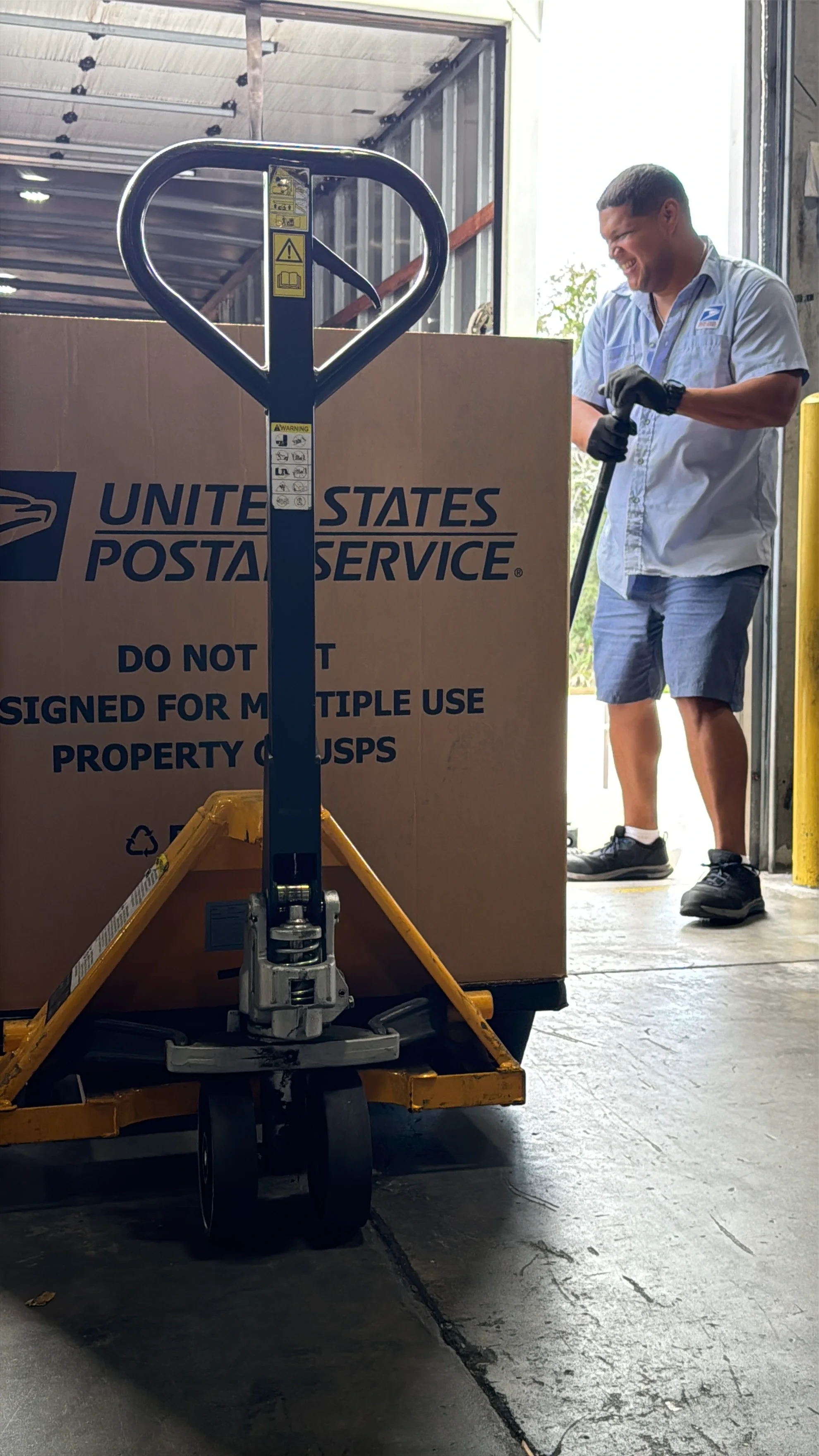 Un hombre con una camiseta de USPS y pantalones cortos operando una carretilla de mano con una gran caja de cartón etiquetada United States Postal Service, dentro de un almacén.