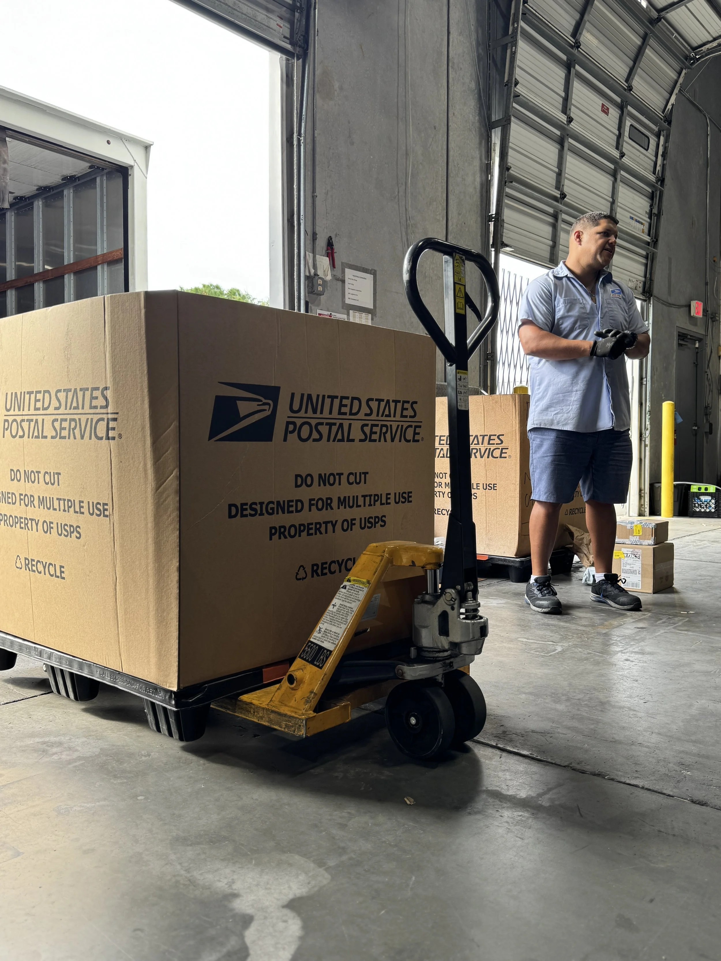 Un hombre con una camisa de trabajo azul claro, pantalones cortos y guantes está de pie dentro de un almacén junto a una gran caja de cartón del United States Postal Service sobre una carretilla. El almacén tiene una gran puerta de garaje abierta y se pueden ver diversas cajas y equipos.