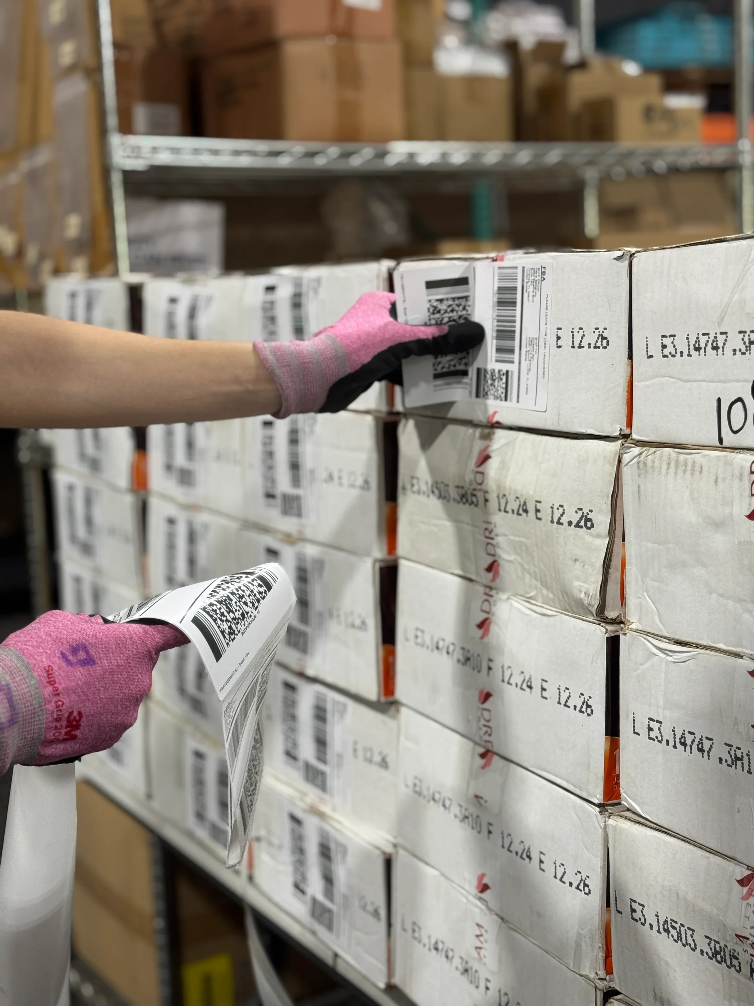 Un trabajador con guantes rosados y negros manipula etiquetas en un almacén con estanterías de cajas apiladas.