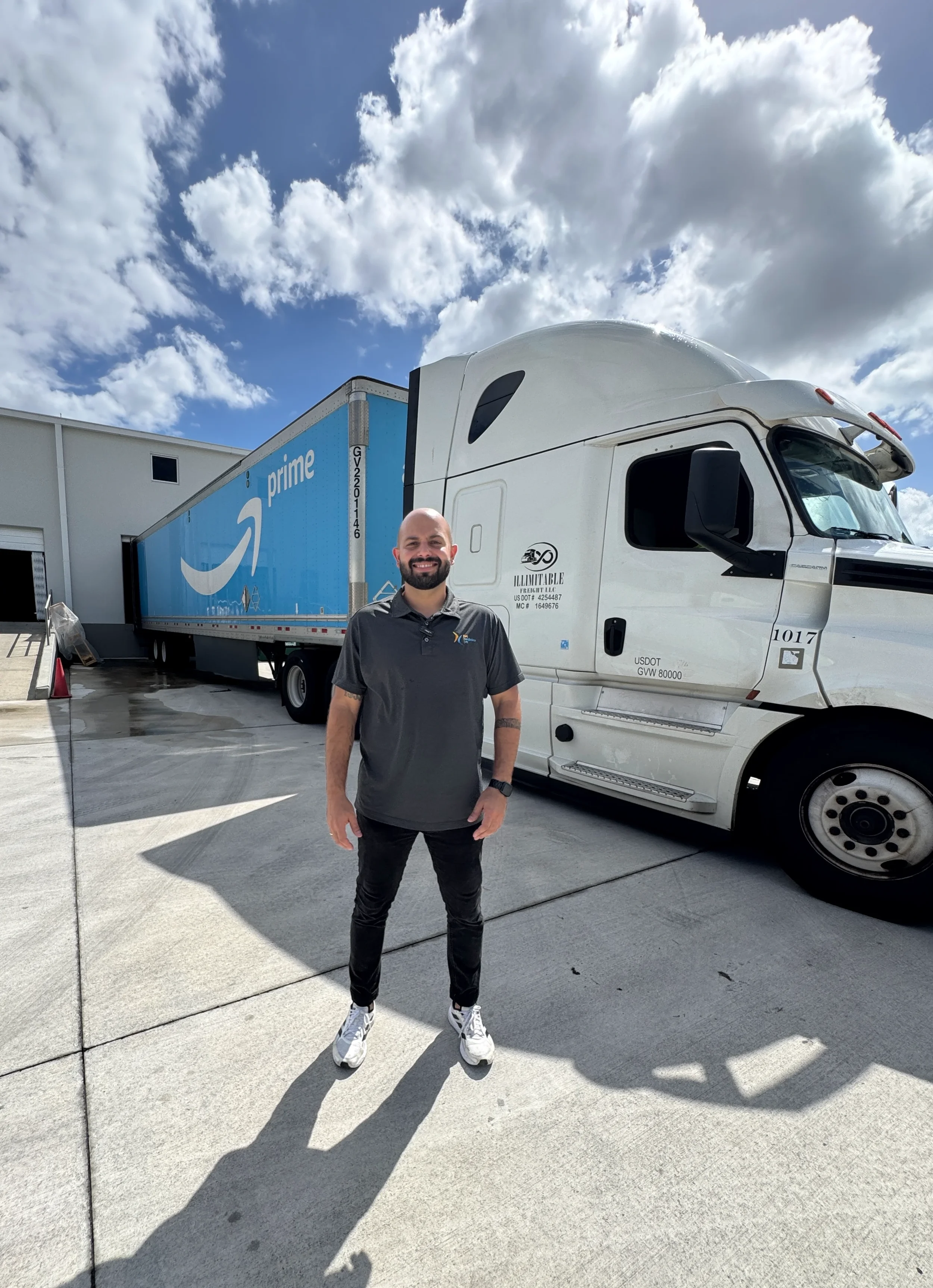 Un hombre con barba está de pie sonriendo frente a un camión semirremolque de Amazon Prime bajo un cielo parcialmente nublado. Lleva una camisa polo gris oscuro, pantalones negros y zapatillas blancas, y parece estar al aire libre en un patio de concreto.